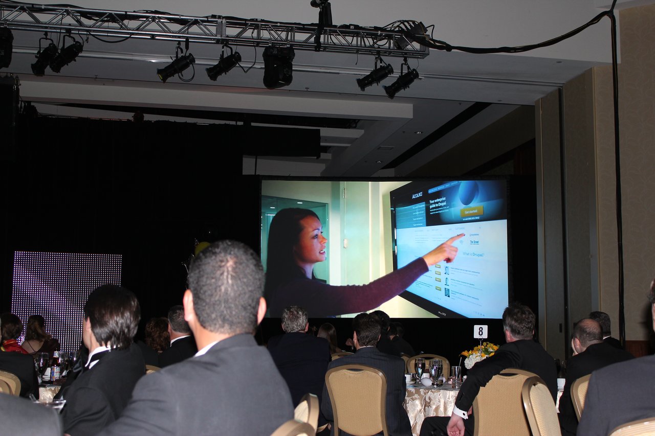 Attendees in formal attire watch a large screen displaying a woman pointing at a webpage during an award banquet.