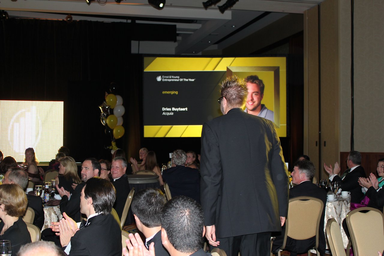 A man in a suit walks toward the stage as the audience claps during an award banquet.