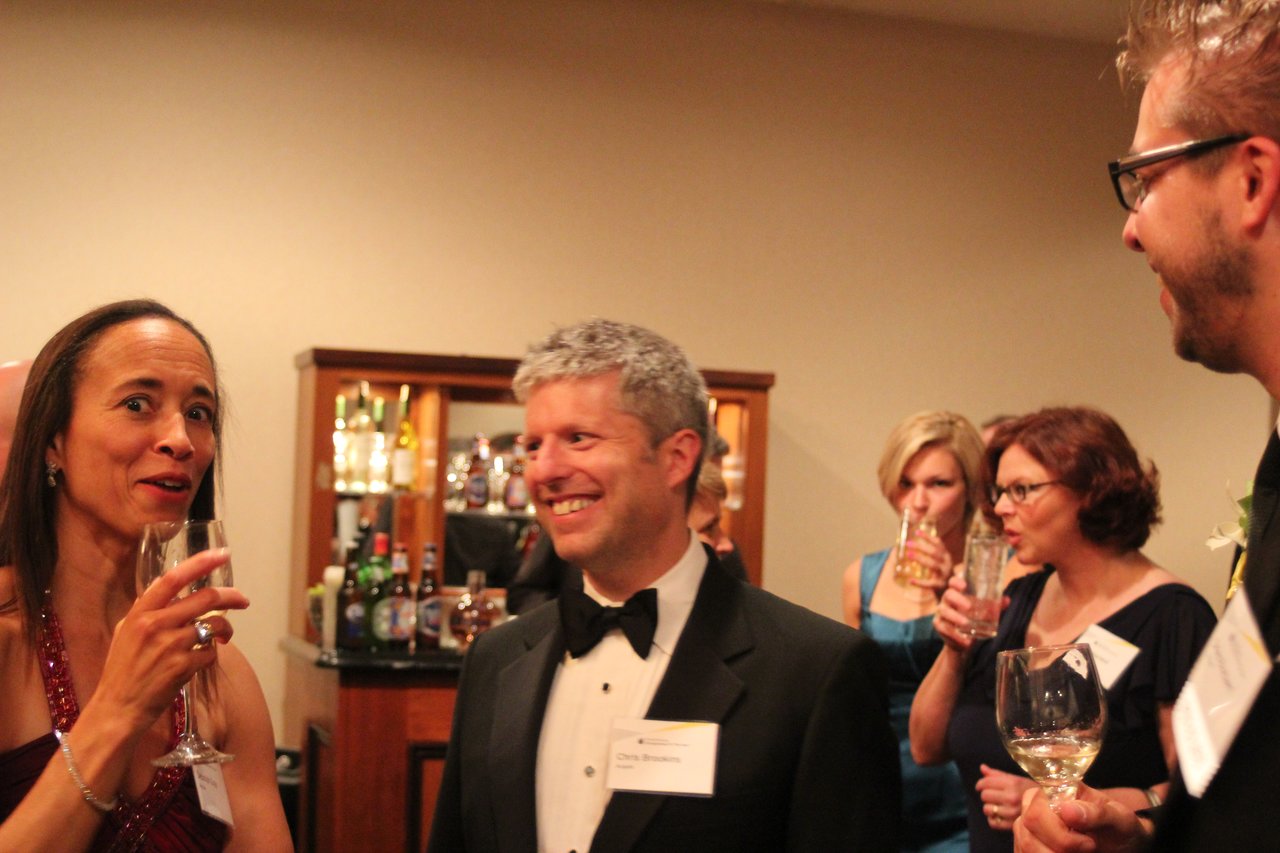 People in formal attire converse and drink wine at an award banquet, with a bar in the background.