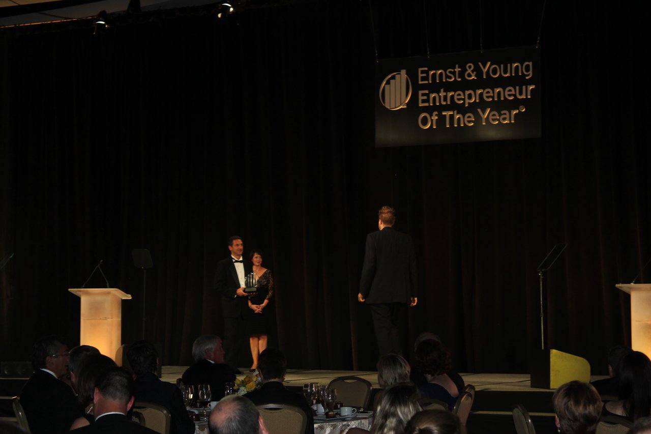 A man in a tuxedo holds an award on stage while another man approaches during a banquet event.