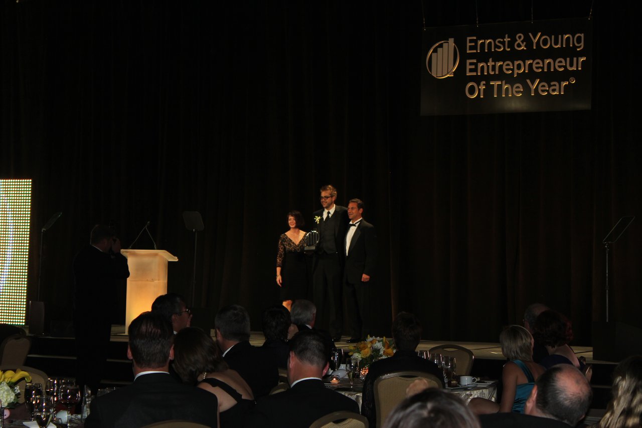 Three people stand on stage at an awards banquet, with one holding a trophy, as the audience watches.