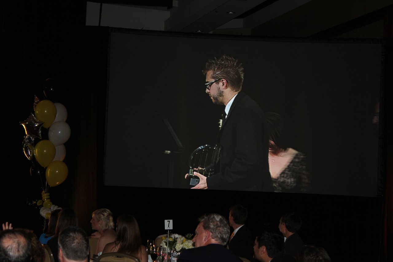 A man in a suit holds an award on stage, displayed on a large screen at a banquet event.