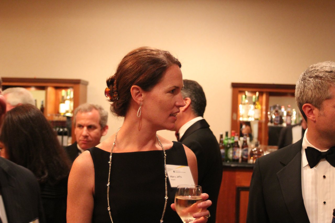 A woman in formal attire holds a glass of wine and talks with others at an award banquet.