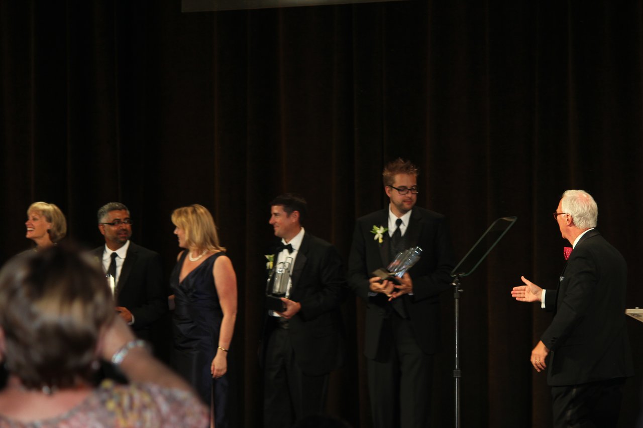 Award recipients in formal attire stand on stage holding trophies while a presenter gestures toward them.