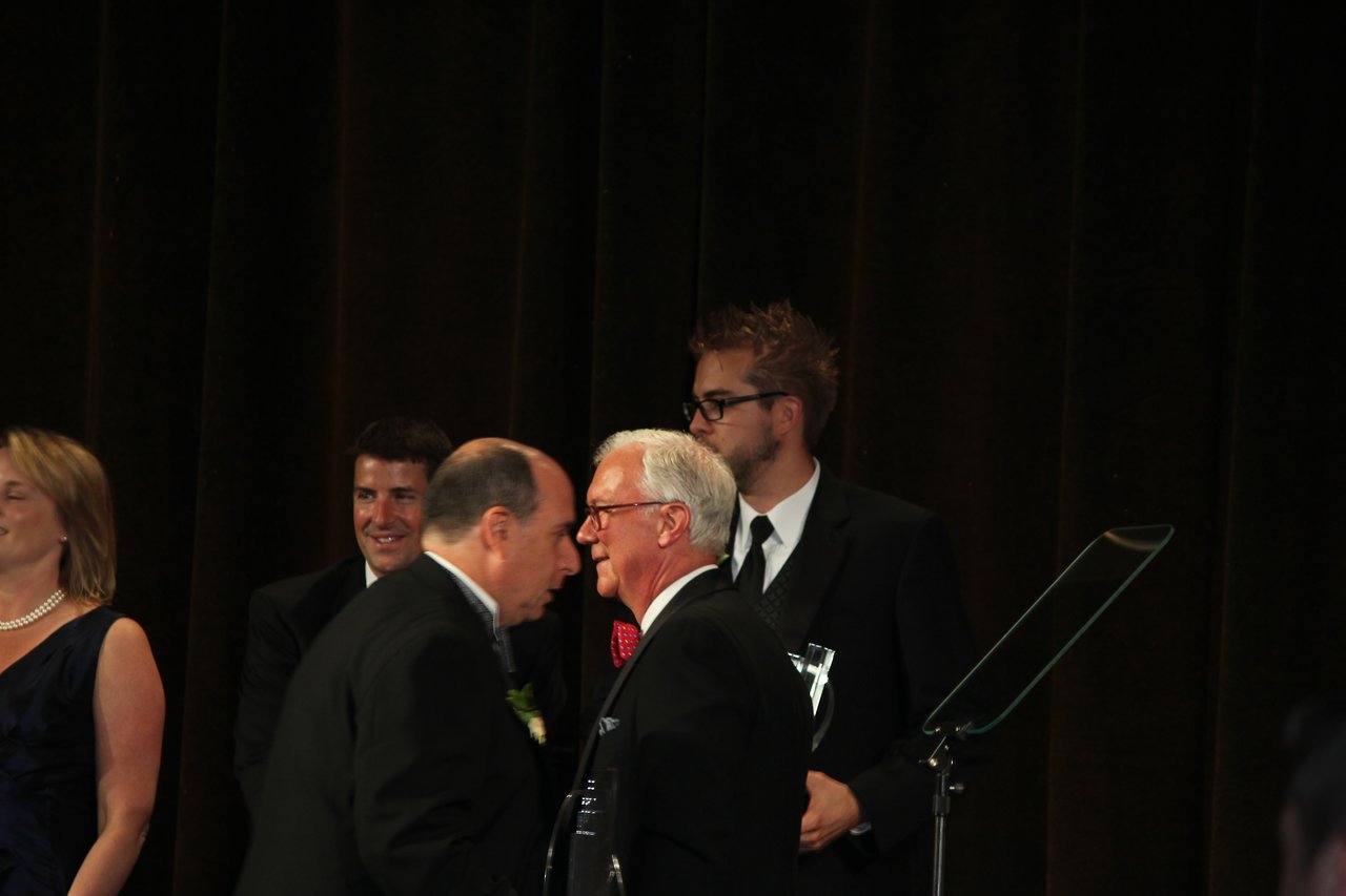 Several people in formal attire stand on stage, with one man handing an award to another during a banquet.