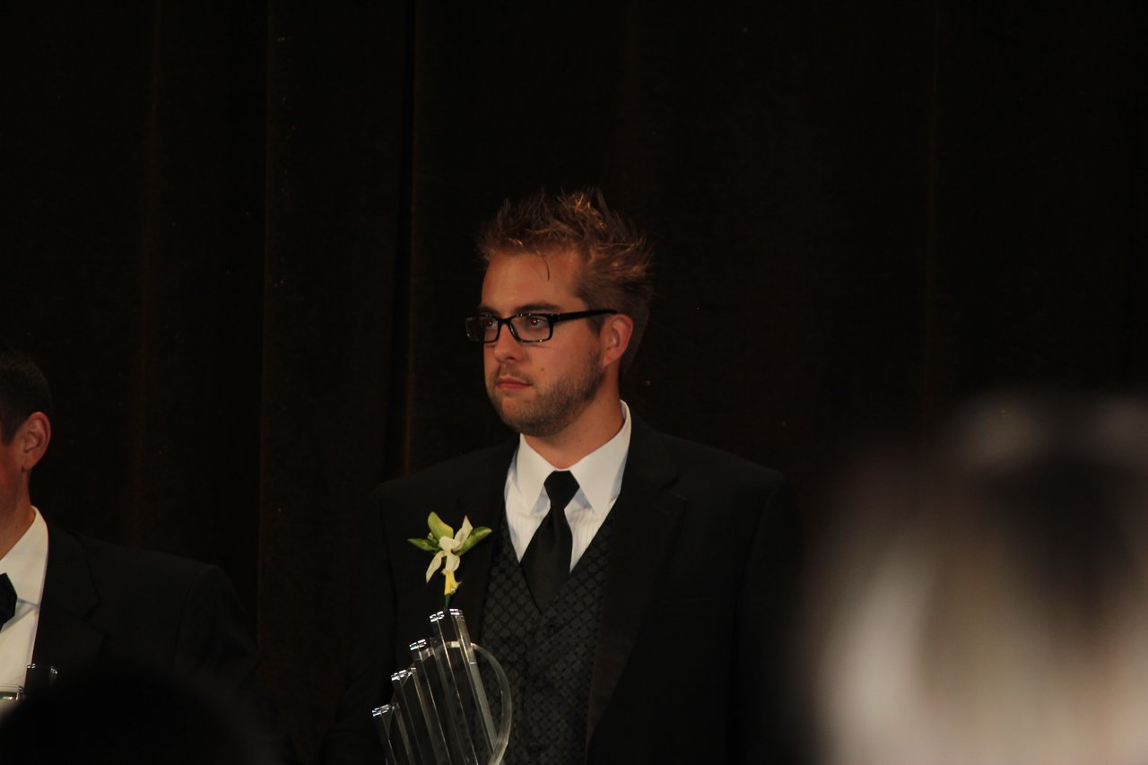 A man in a suit and glasses stands on stage holding an award at a banquet event.