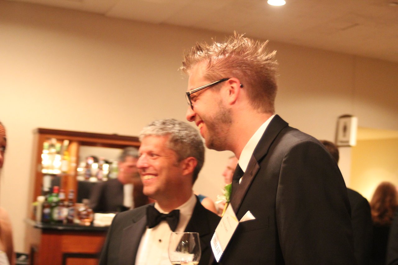 Two men in tuxedos smile and converse at an award banquet, with a bar and other guests in the background.