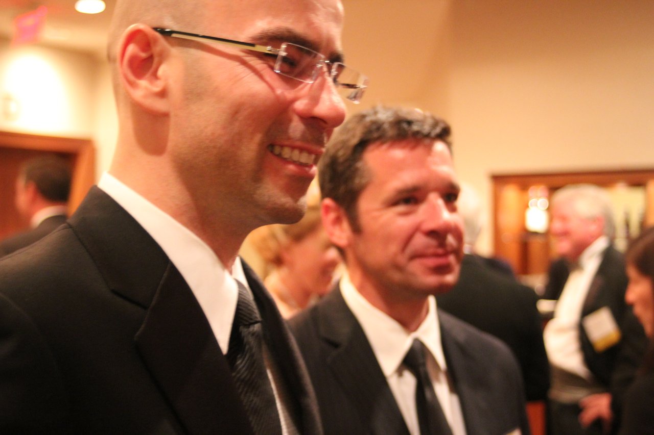 Two men in suits smiling and conversing at an award banquet, with other attendees in the background.