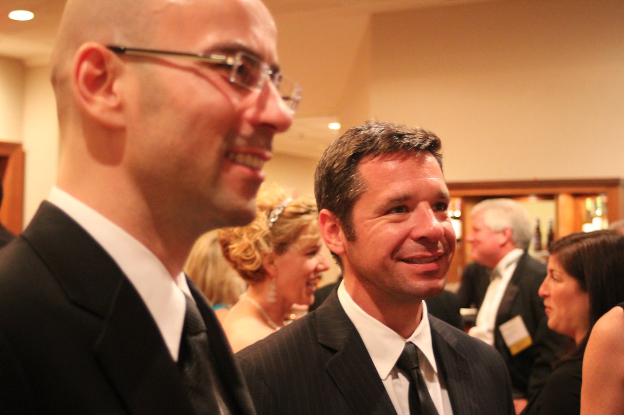 Two men in suits smiling and conversing at an award banquet, with other attendees socializing in the background.