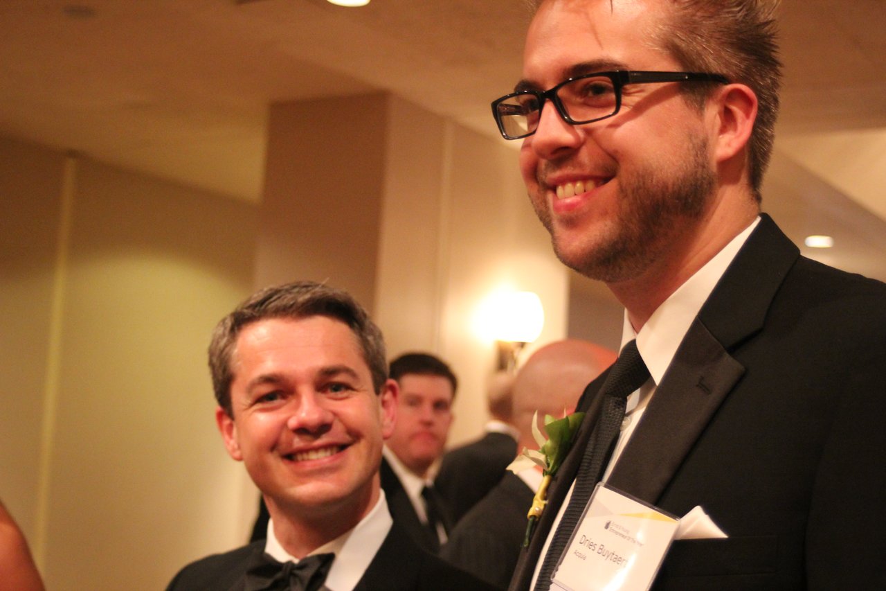 Two men in tuxedos smiling at an award banquet, one wearing a name tag and boutonniere.