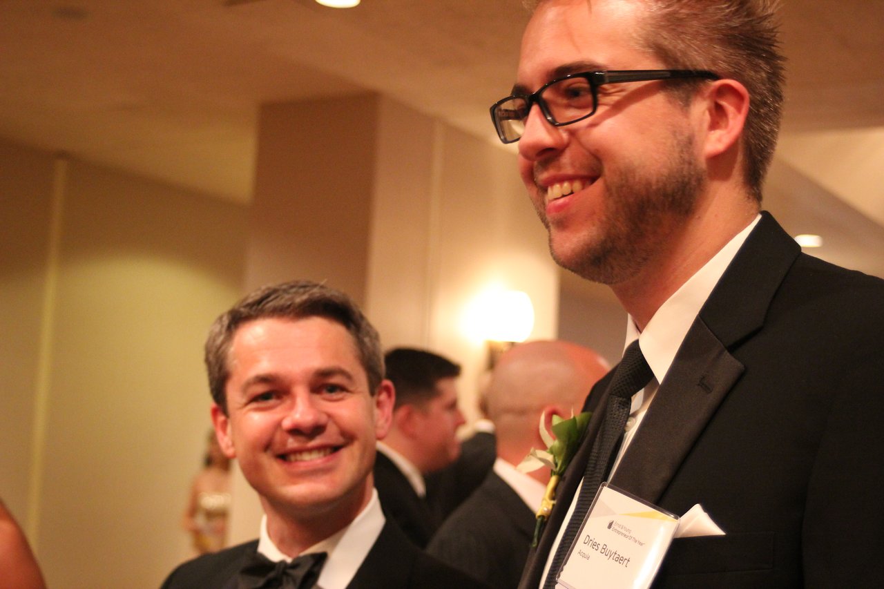 Two men in tuxedos smiling and talking at an award banquet, with other attendees in the background.