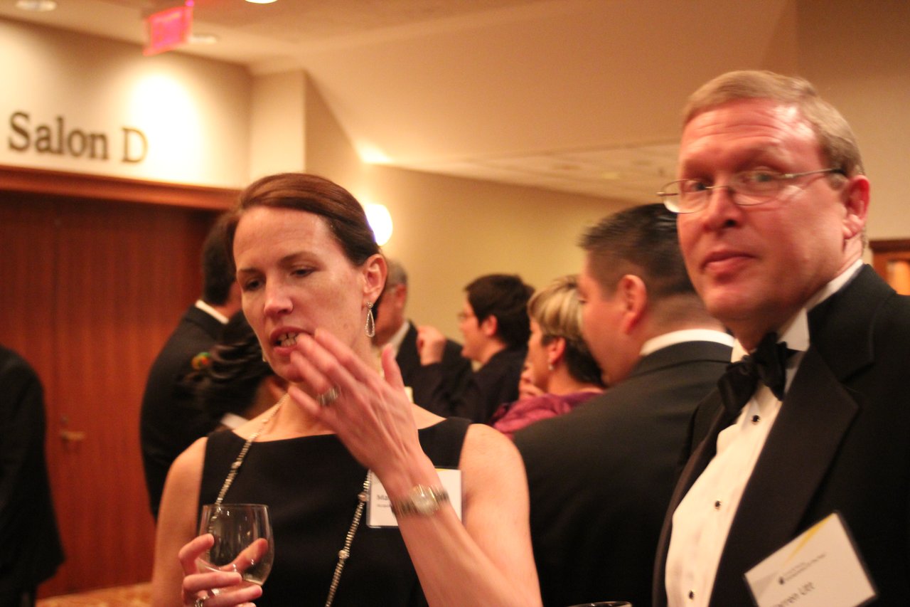 Two formally dressed people converse at an award banquet, surrounded by other attendees in a crowded reception area.