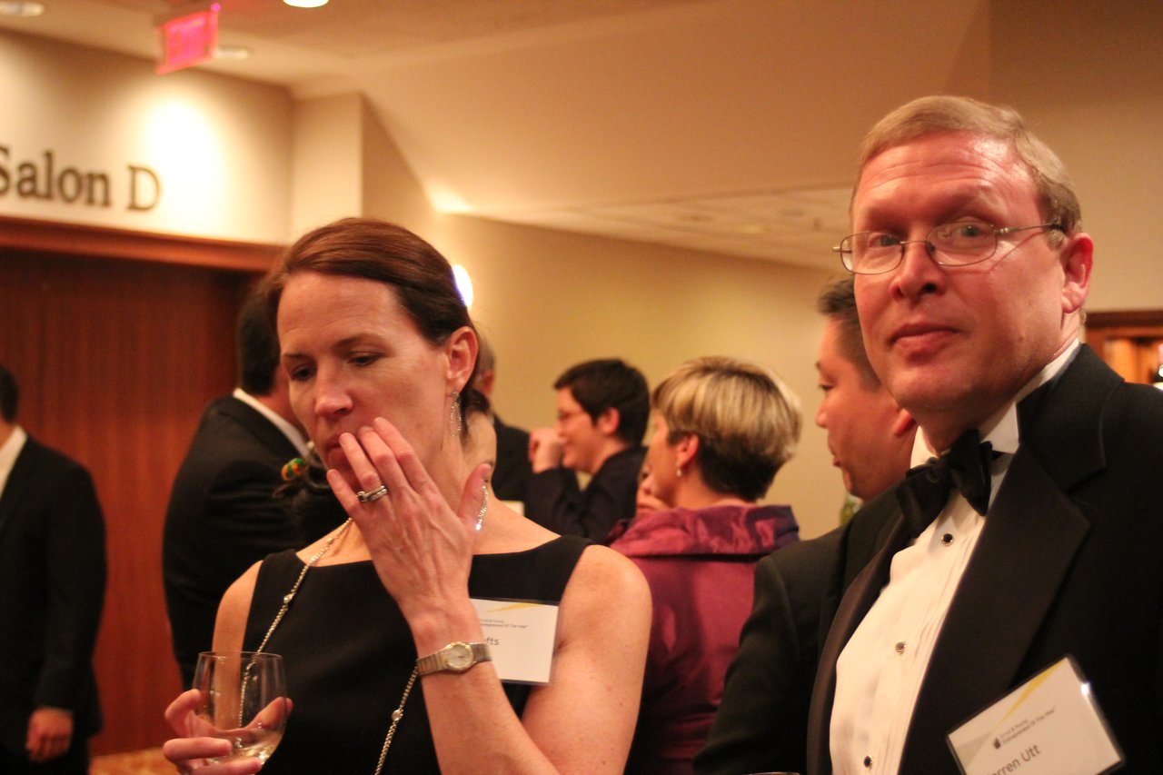 A man in a tuxedo and a woman with a drink attend a formal award banquet, surrounded by guests.