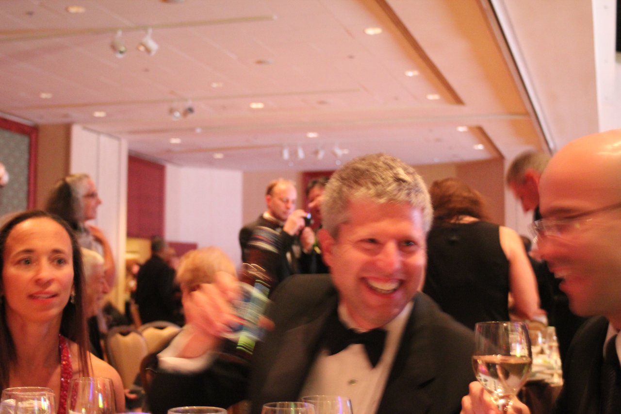 People in formal attire are seated at a banquet table, smiling and toasting with drinks in a lively setting.