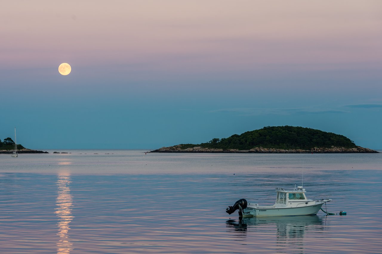 A full moon reflects on calm ocean water with a small boat floating nearby and an island in the background.