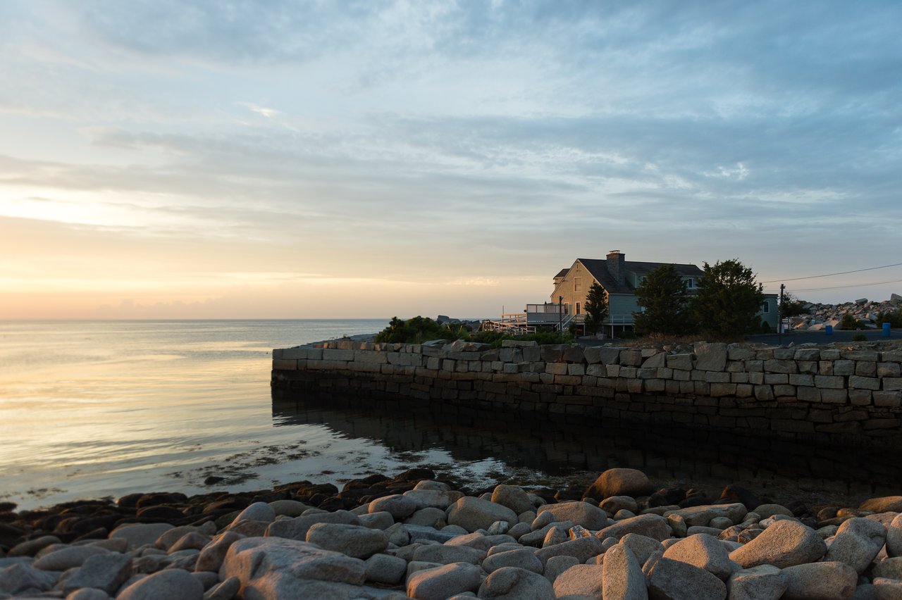 A coastal scene at sunset with a stone seawall, rocky shore, and a house overlooking calm waters.