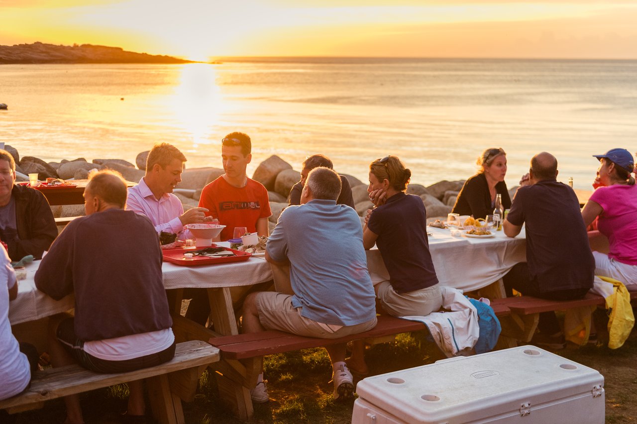 A group of people sitting at picnic tables by the water, eating lobster and talking during sunset.