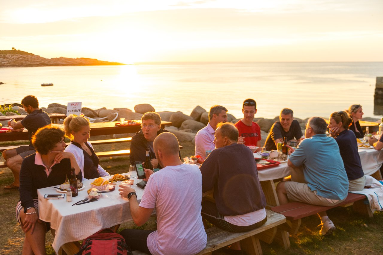 A group of people enjoying a lobster dinner at outdoor tables by the water during sunset.