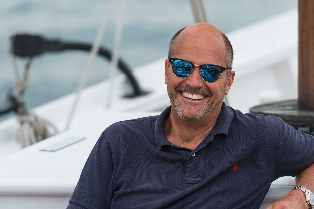 A man wearing sunglasses and a navy polo shirt smiles while relaxing on a sailboat.
