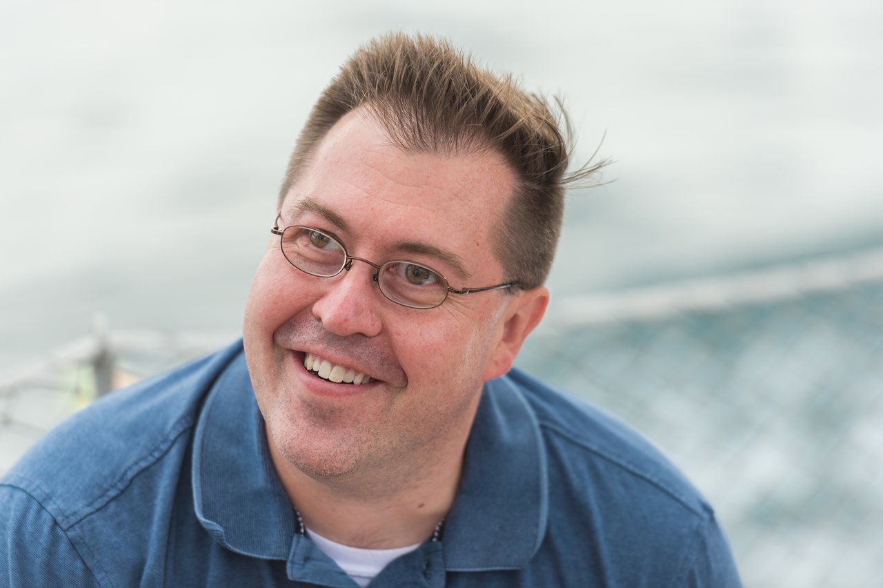 A smiling person wearing glasses and a blue shirt on a boat with water in the background.