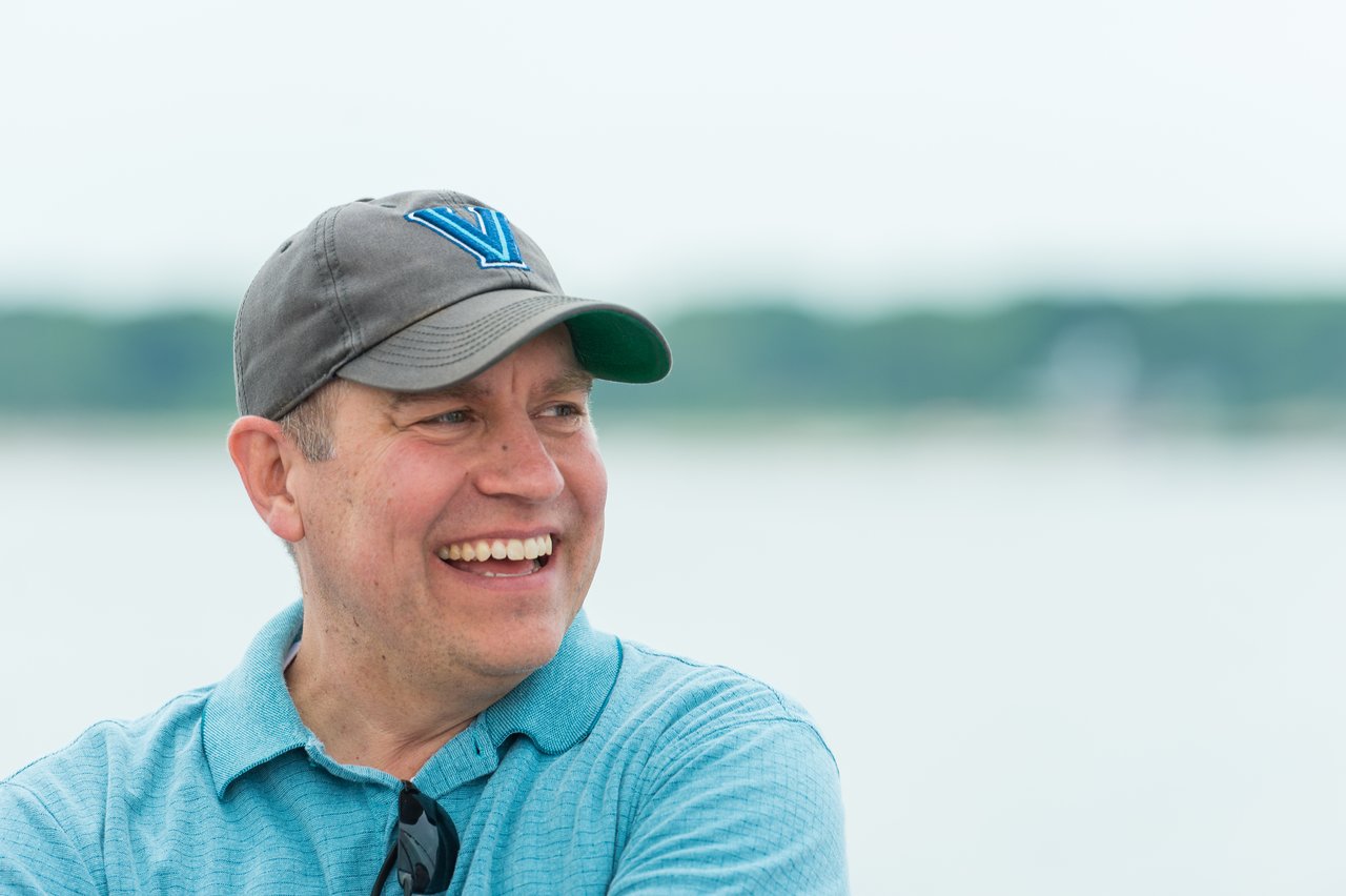A man in a cap and polo shirt smiles while looking to the side during a sailing trip.