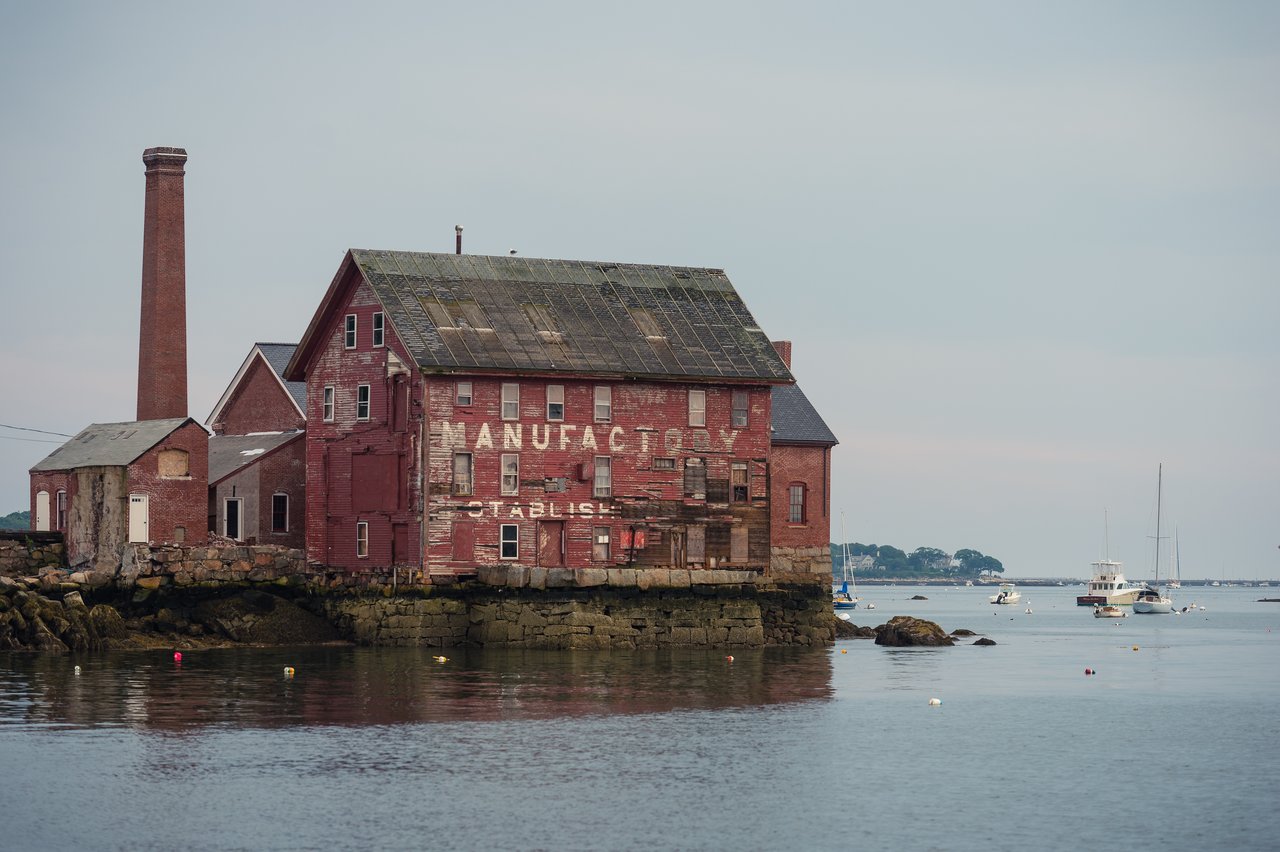 An old red brick building by the water with sailboats and motorboats anchored nearby.
