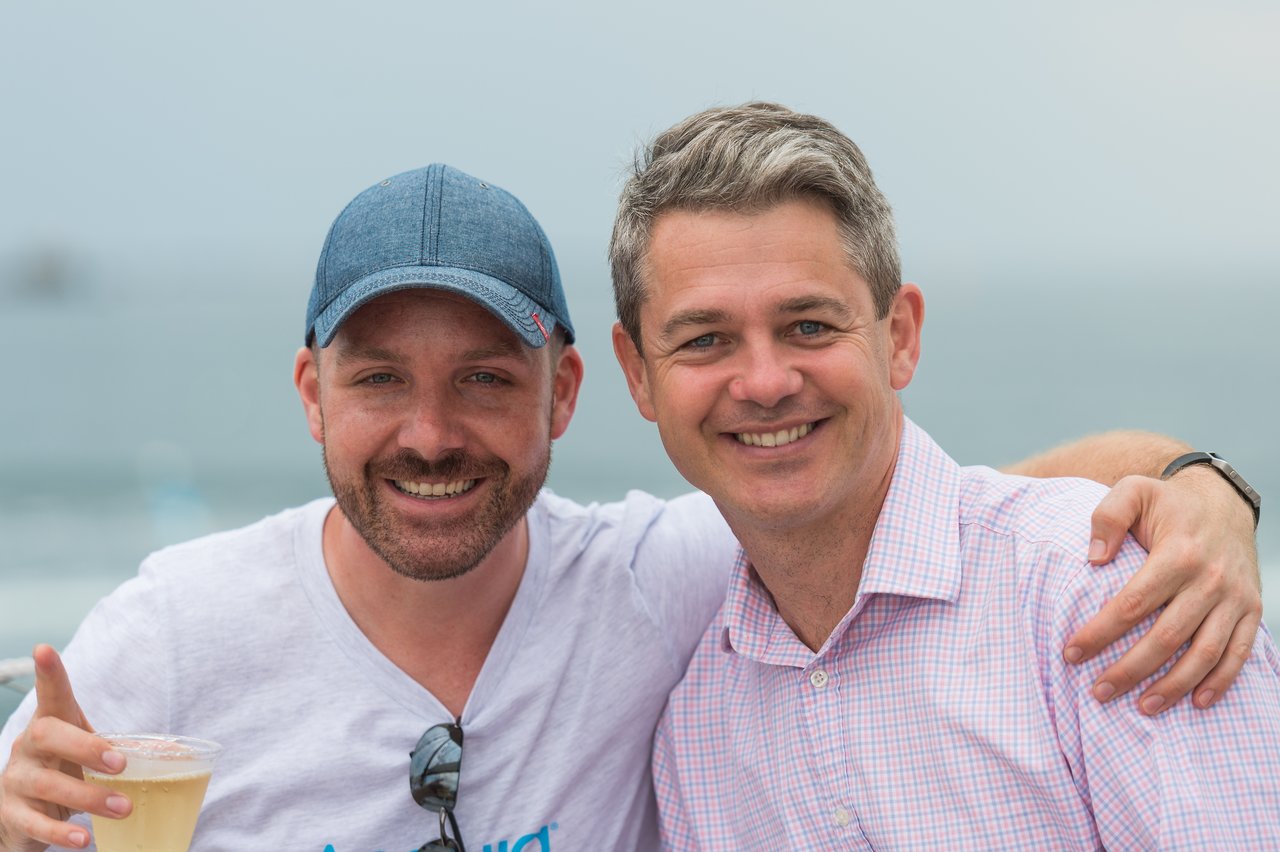 Two men smiling, one wearing a cap and holding a drink, with an arm around the other's shoulder.