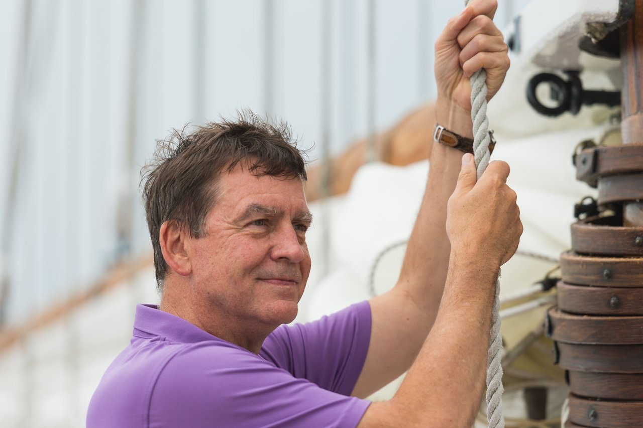 A man in a purple shirt holds a rope while adjusting sailing equipment on a boat.