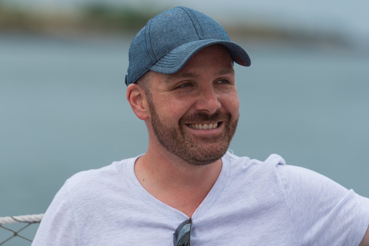 A man wearing a blue cap and white shirt smiles while sailing on the water.