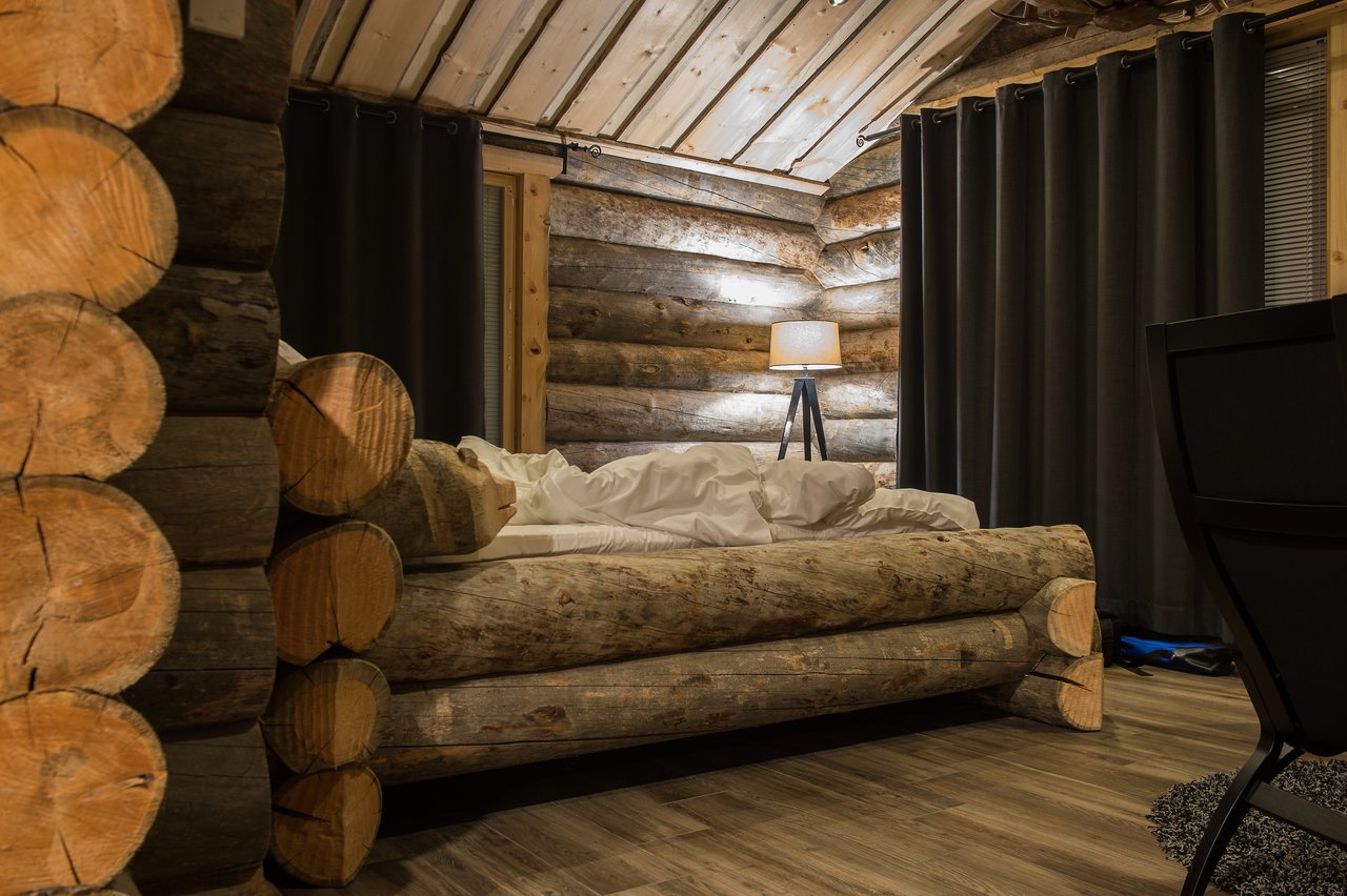 A rustic log cabin bed with white bedding, surrounded by wooden walls and a lit bedside lamp.