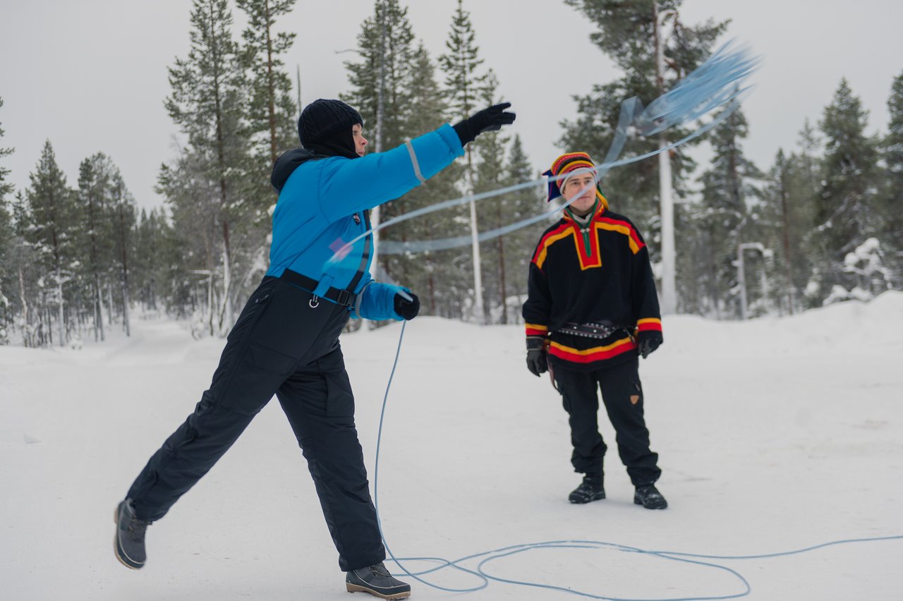 A person in winter clothing practices lassoing, while another in traditional attire observes in a snowy outdoor setting.