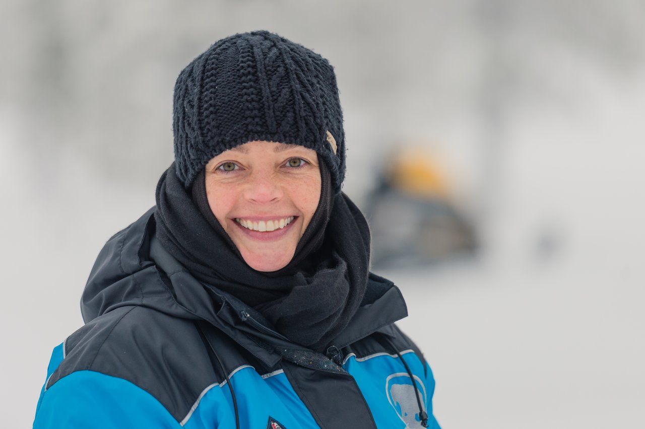 A person in winter clothing smiles outdoors in a snowy setting.