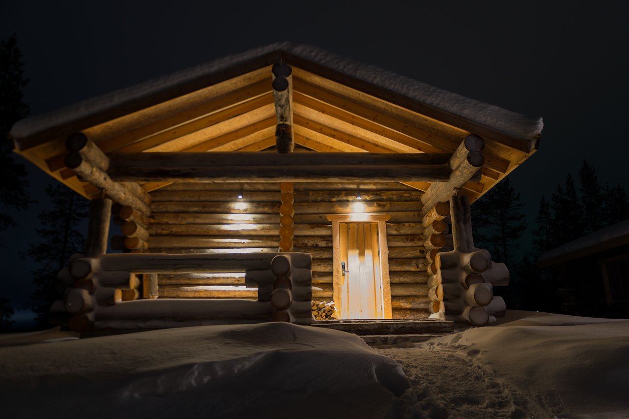 A wooden log cabin with lights on, surrounded by snow at night, with a path leading to the door.
