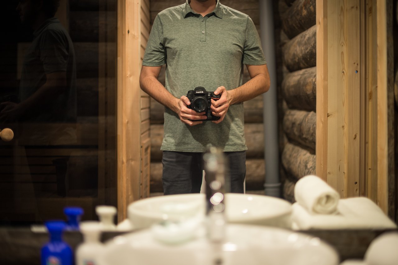A person in a green shirt takes a mirror selfie with a camera in a log cabin bathroom.