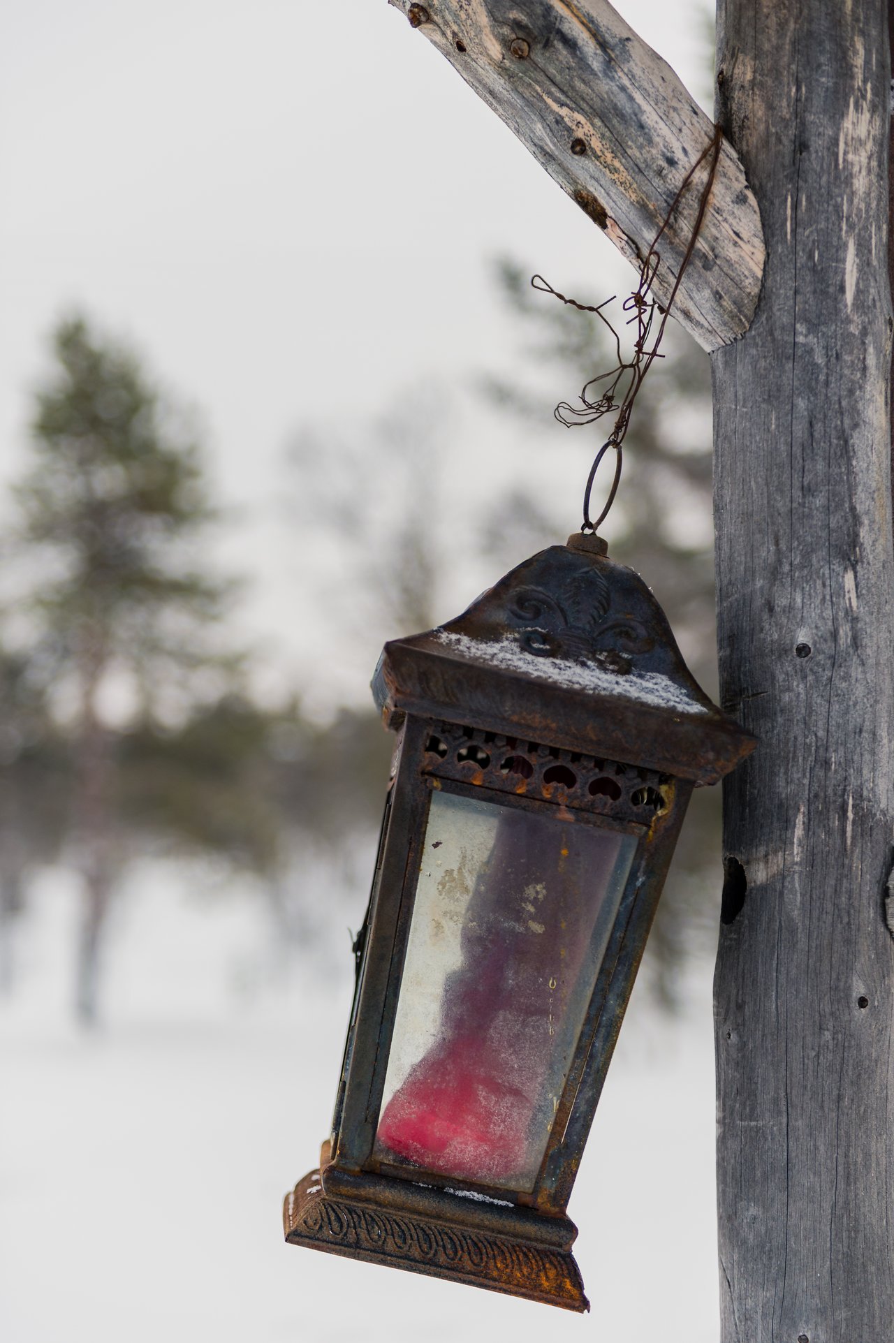 A rusty outdoor lantern with a red candle inside, hanging from a wooden post in a snowy setting.