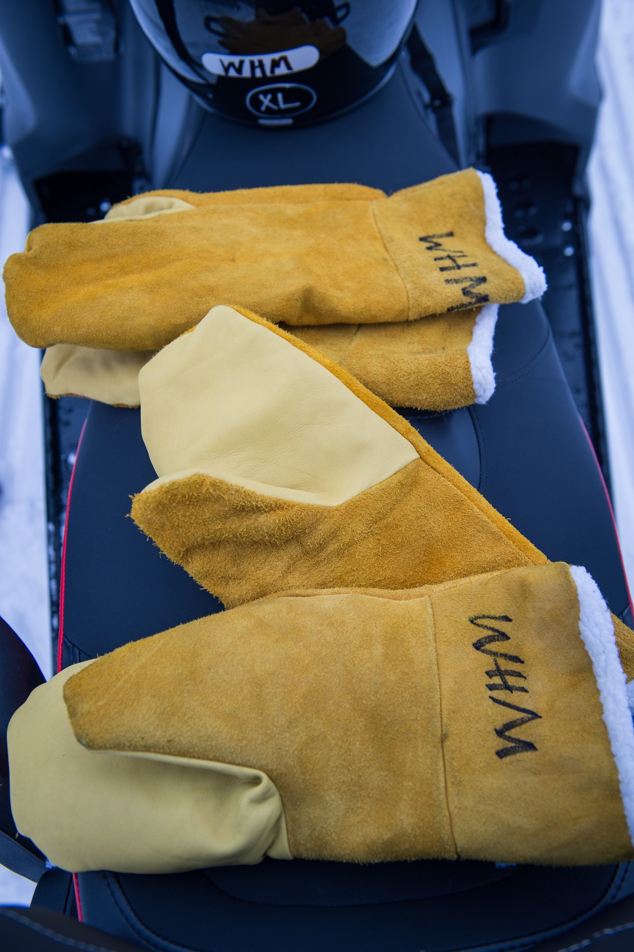 A pair of thick, insulated mittens with suede and leather material rest on a black seat, with "WHM" marked.