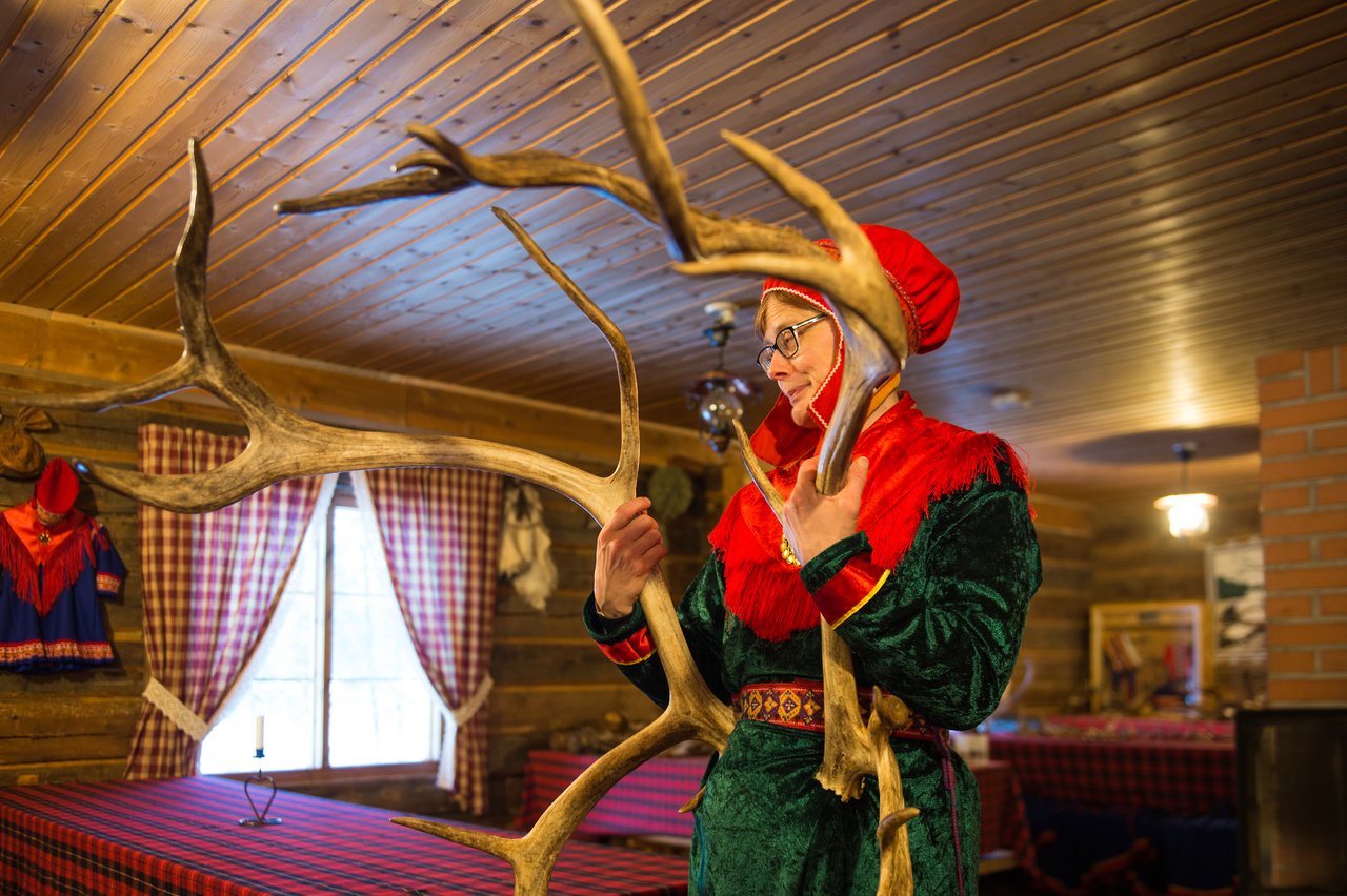 A person in traditional clothing holds large reindeer antlers inside a wooden cabin with checkered tablecloths and curtains.