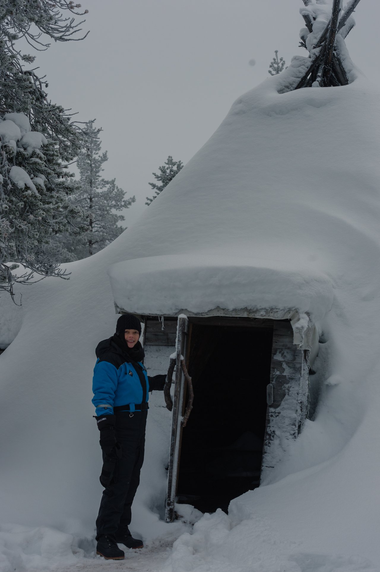 A person in winter clothing stands outside a snow-covered wooden hut, holding the door open and smiling.