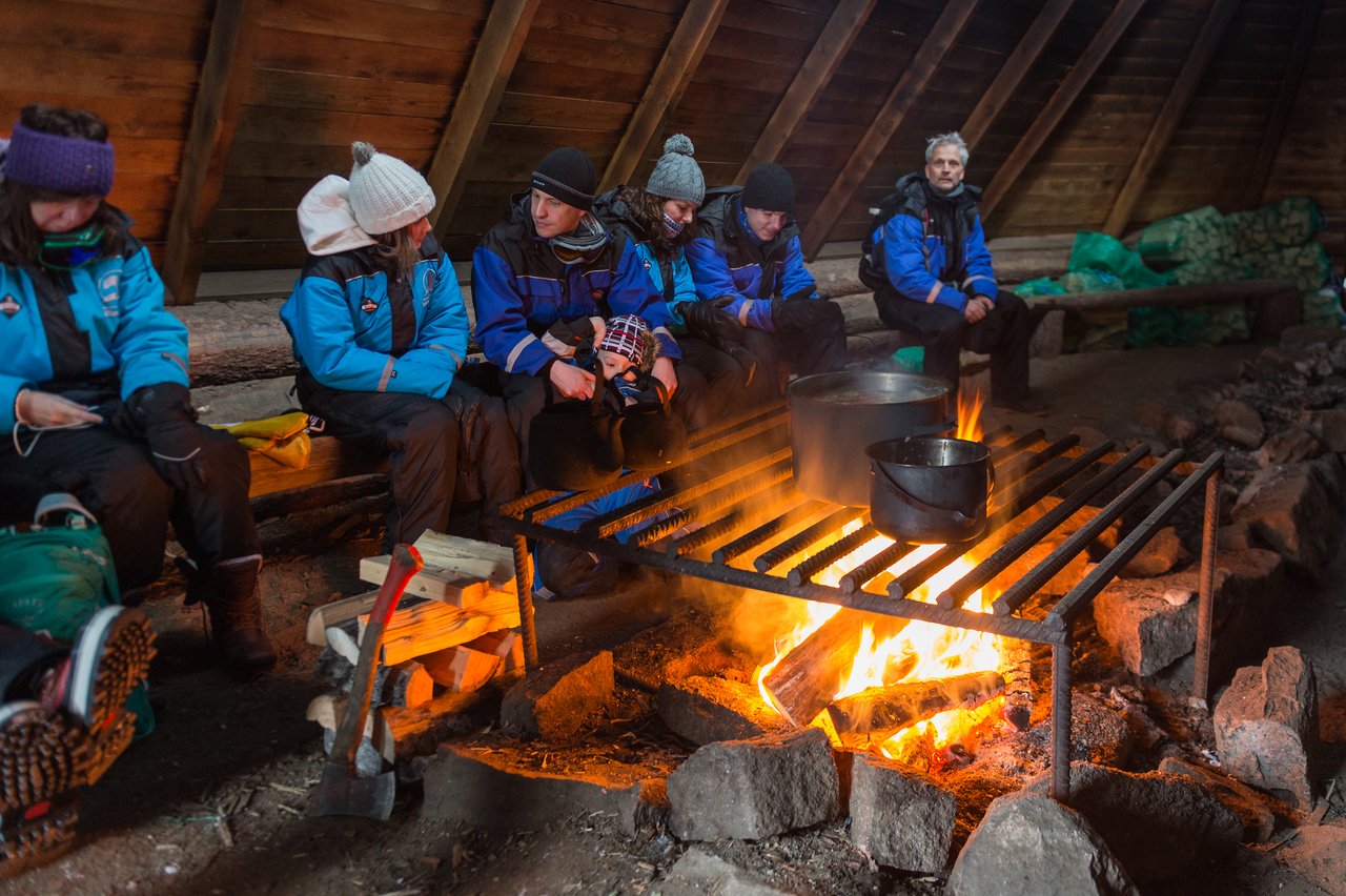A group of people in winter clothing sit by a fire while pots of soup cook on a grill.