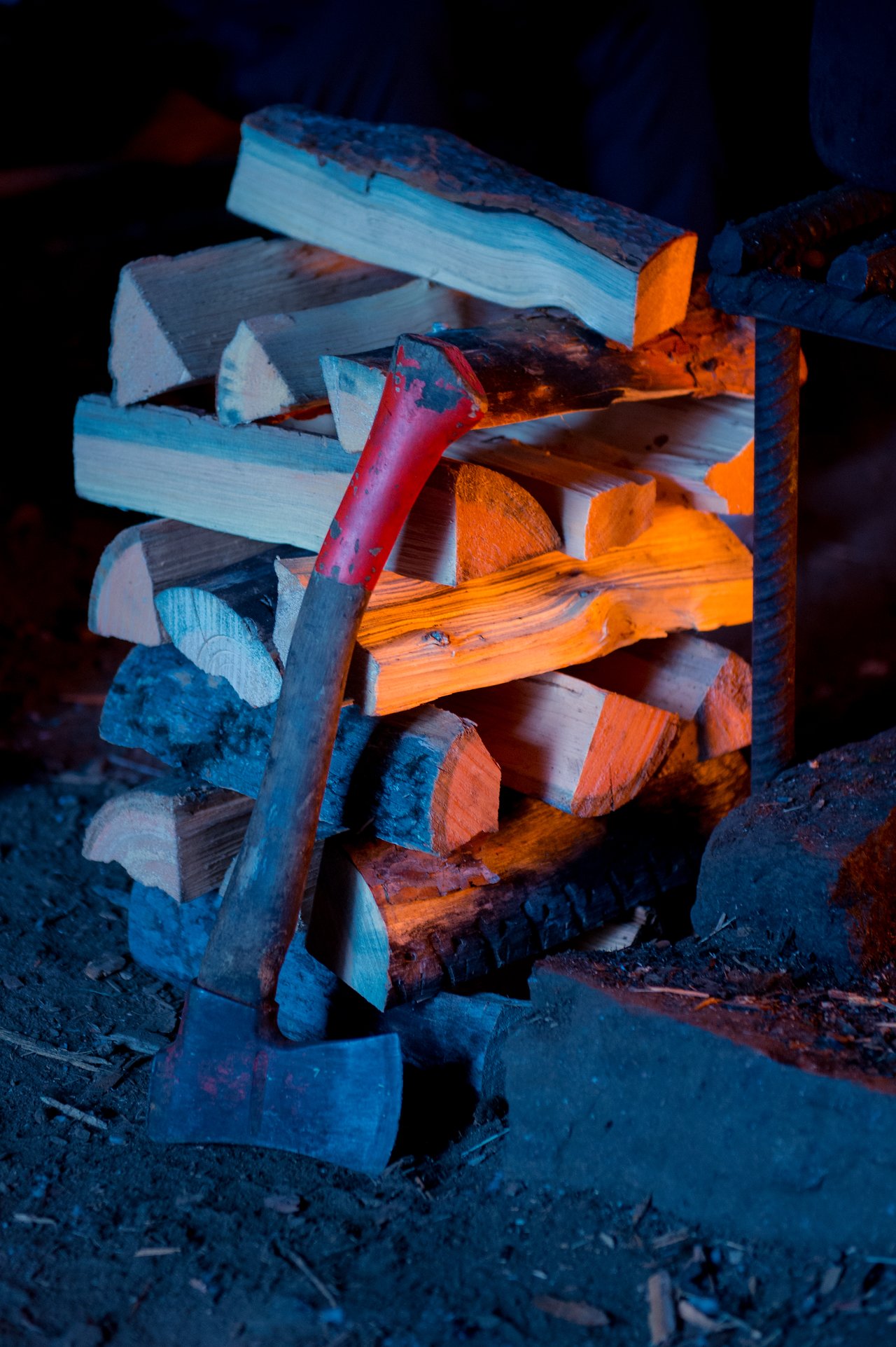A stack of firewood with an axe leaning against it, illuminated by warm firelight in a dim setting.
