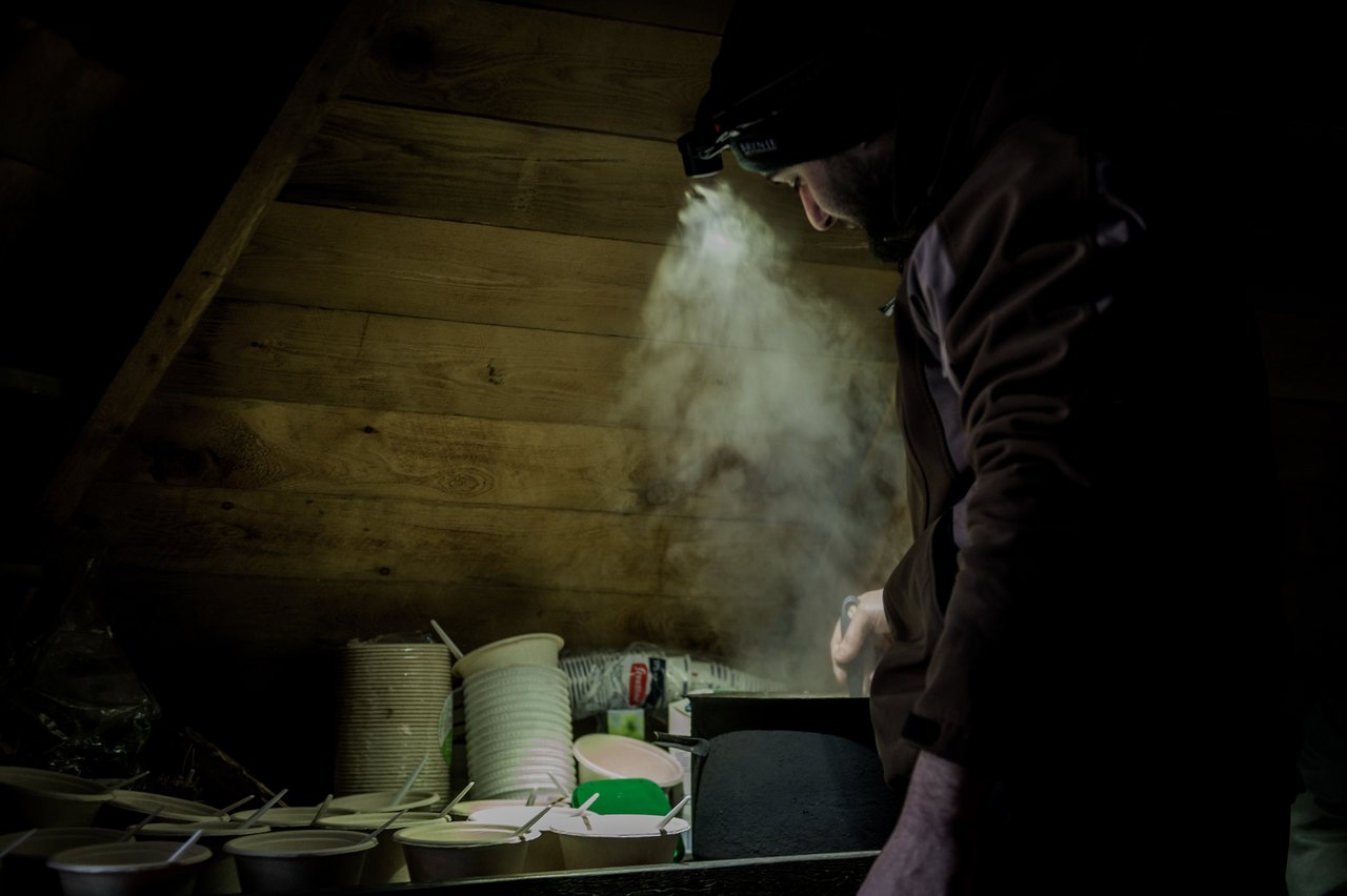 A person in a dark jacket stirs a steaming pot of salmon soup, with bowls and spoons ready nearby.