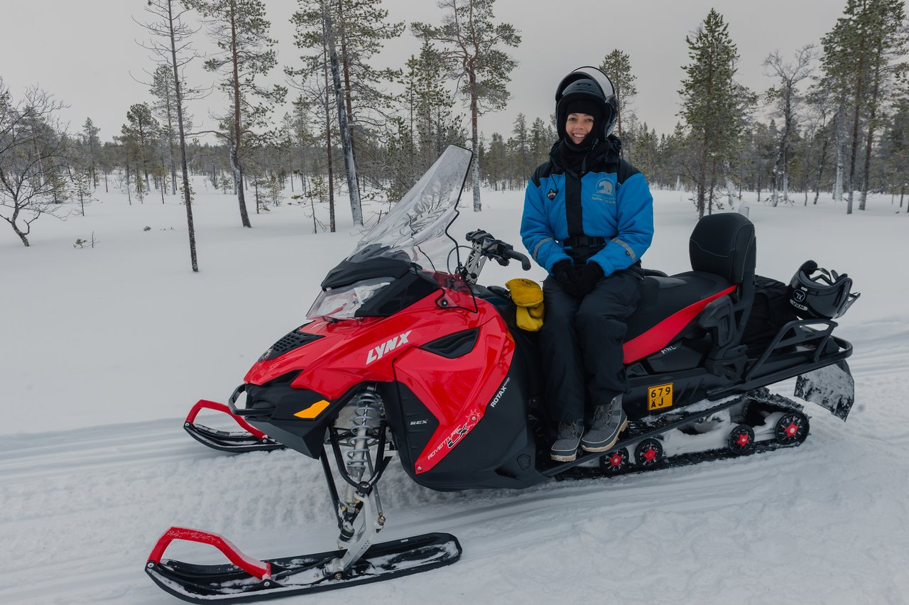 A person in winter gear sits on a red snowmobile in a snowy forest during a Lapland snowmobiling trip.