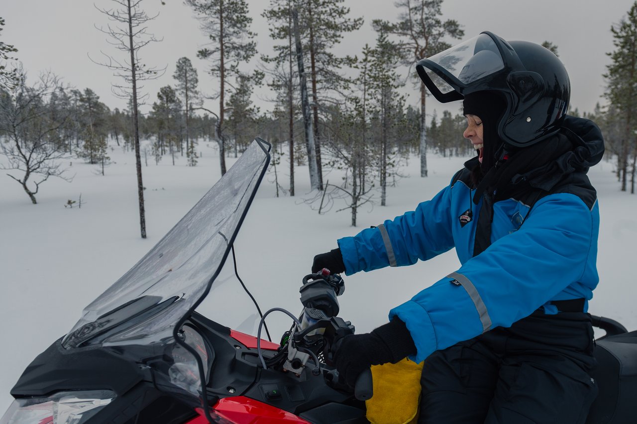 A person wearing a helmet and winter gear rides a snowmobile through a snowy forest in Lapland.