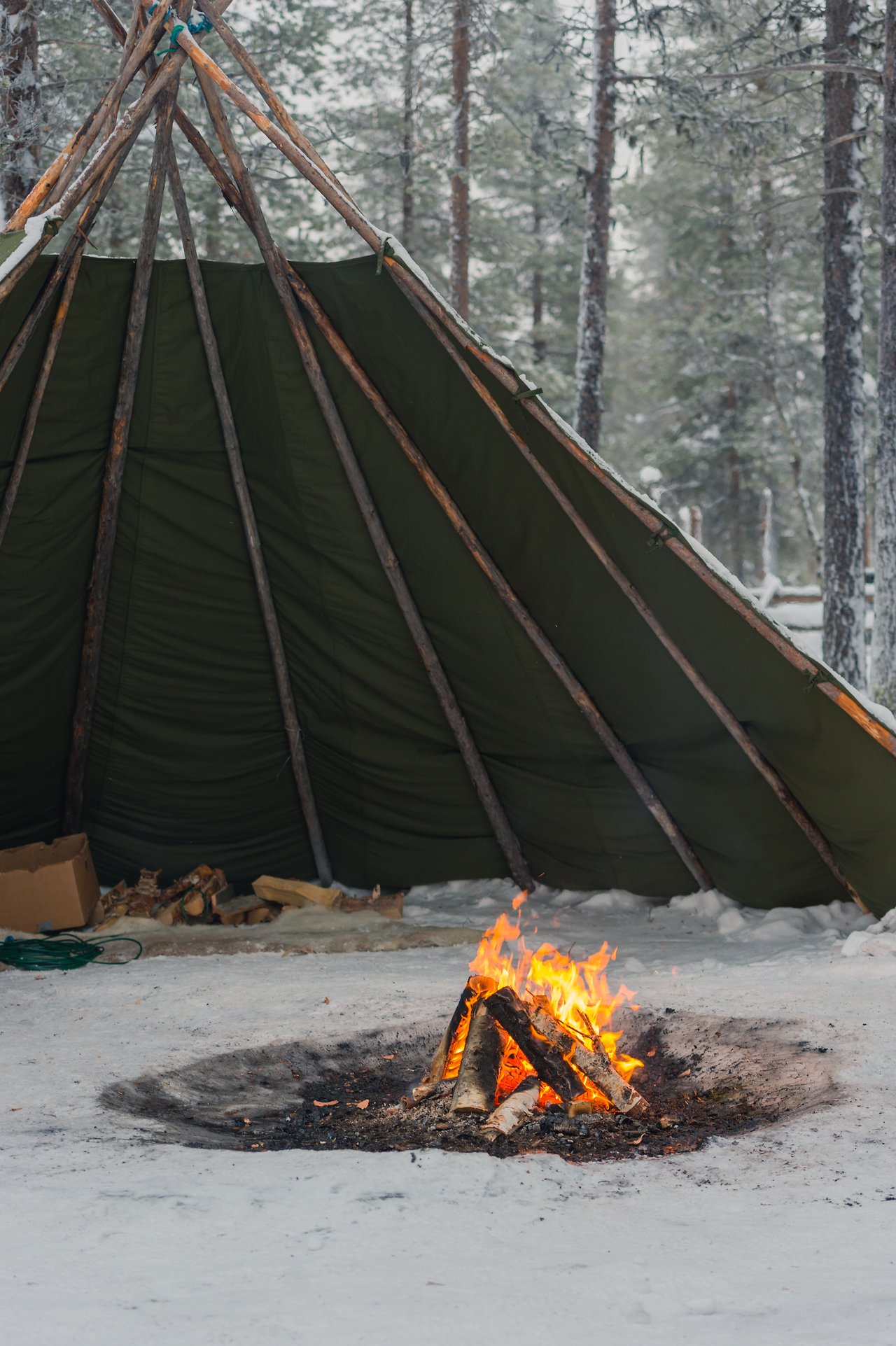 A small campfire burns on snowy ground in front of a partially open tent made of wooden poles and fabric.