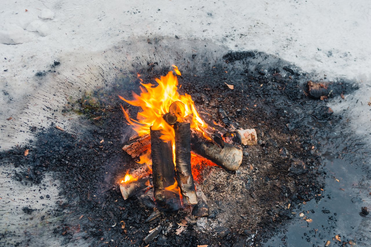 A small campfire burns with wooden logs on snowy ground, providing warmth in cold outdoor surroundings.