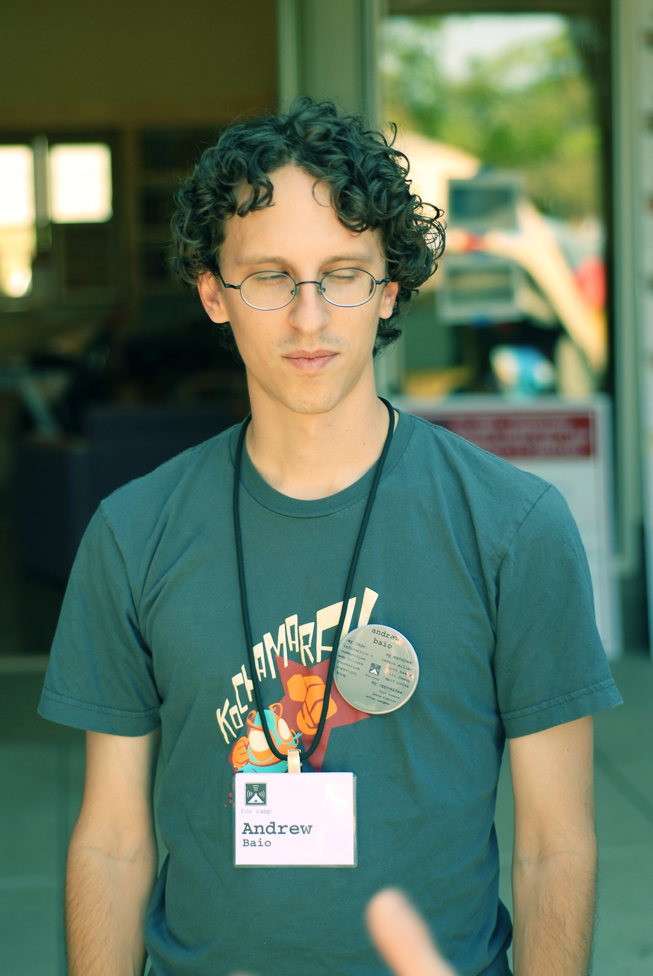 A person wearing glasses and a name tag stands outside, looking downward with a neutral expression.