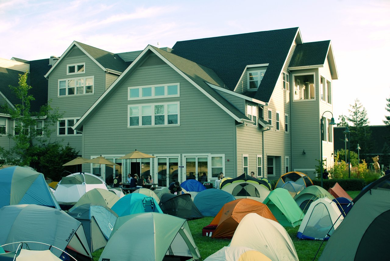 A grassy area filled with tents in front of a large house, with people gathered near tables under umbrellas.