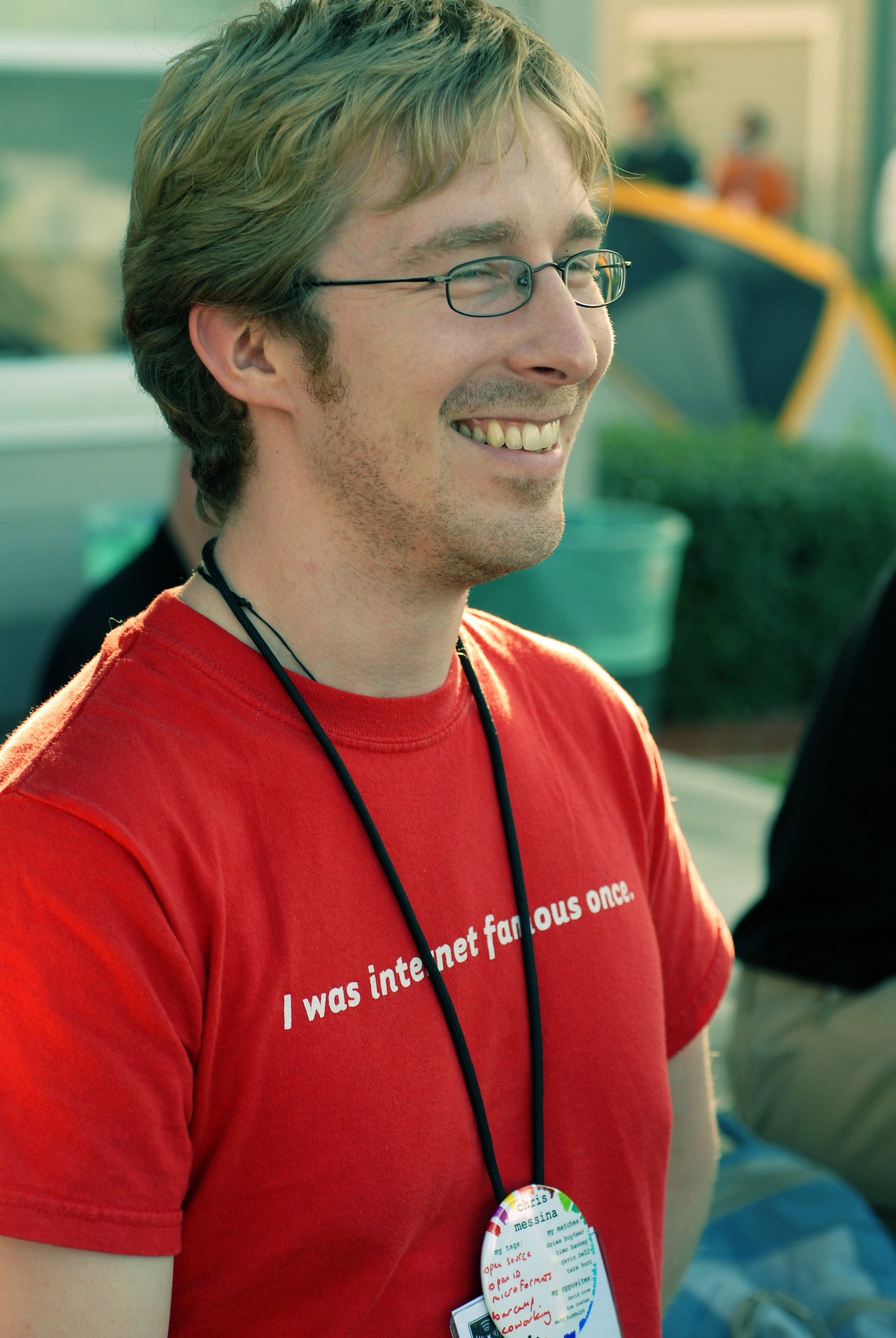 A man wearing glasses and a red t-shirt smiles while attending an outdoor event.
