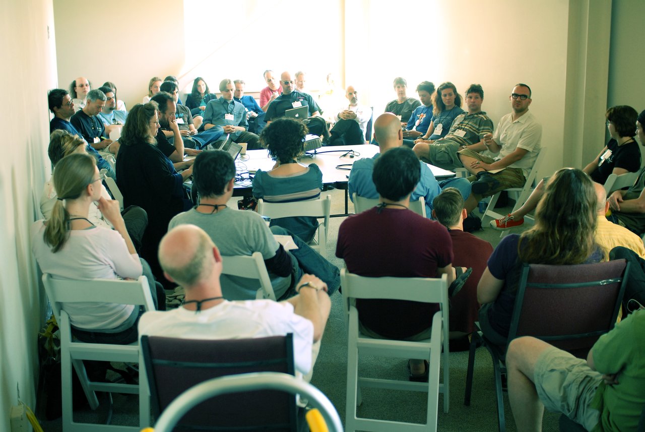 A group of people sit in a circle, attentively listening to a speaker in a discussion setting.