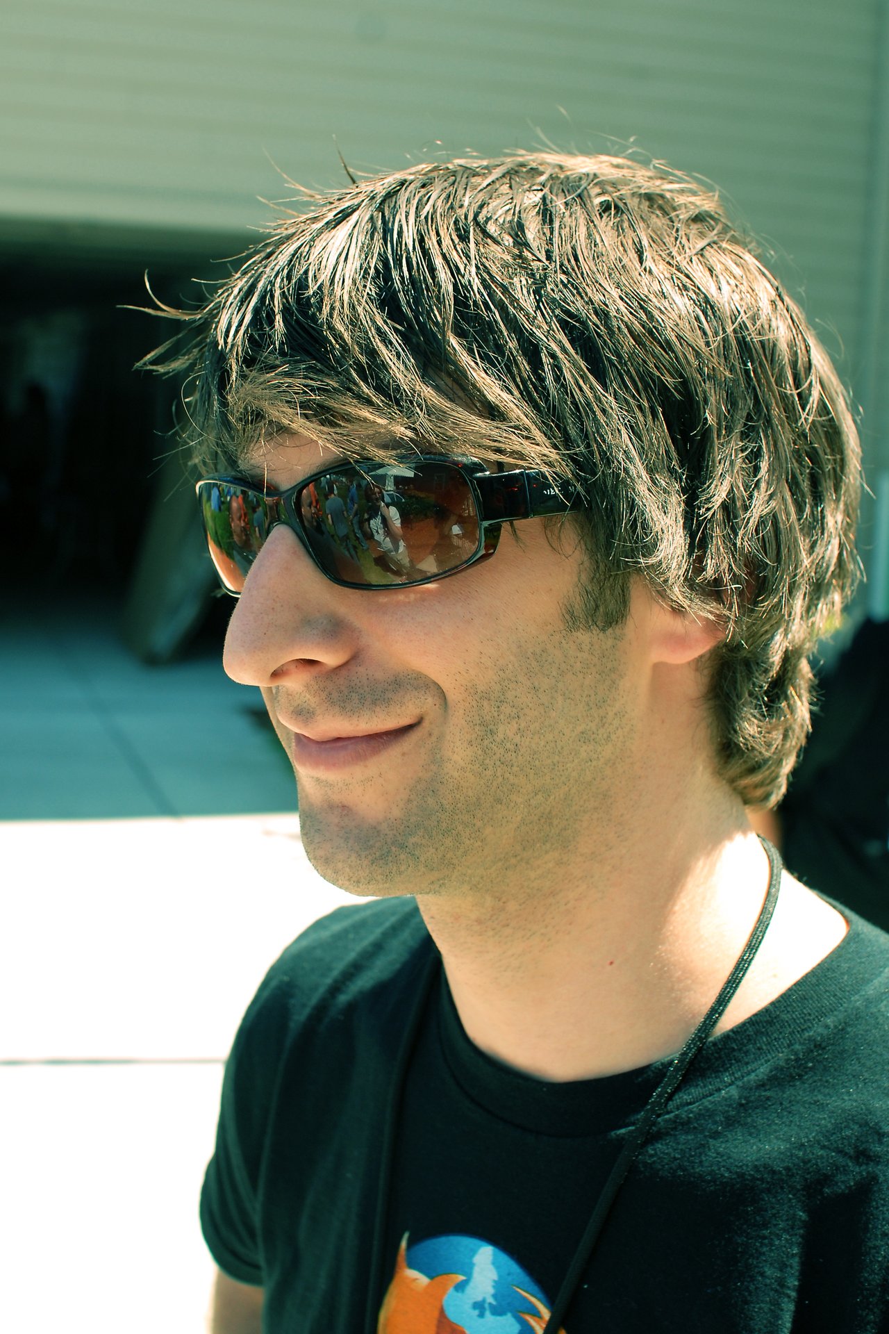 A man wearing sunglasses and a black T-shirt smiles while standing outdoors in bright sunlight.